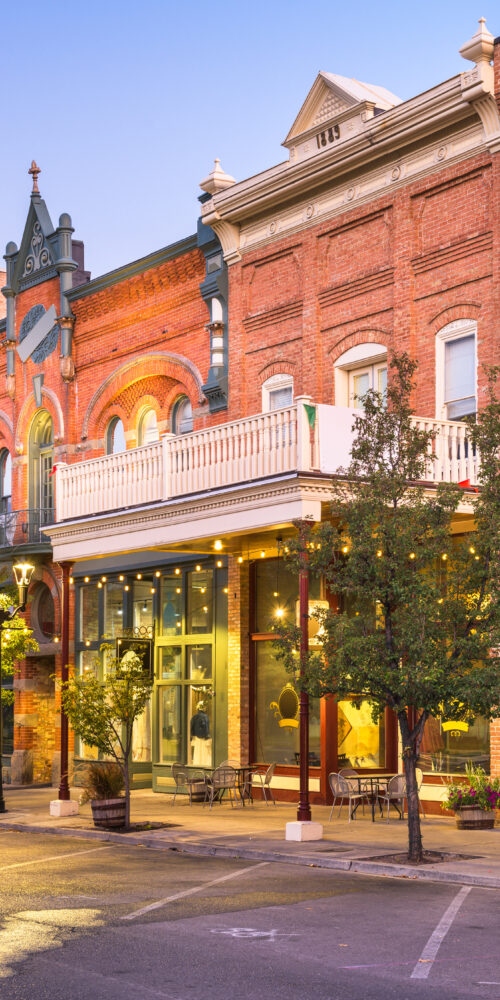 Row of historic brick storefronts with arched windows and outdoor seating along a tree-lined street at dusk.
