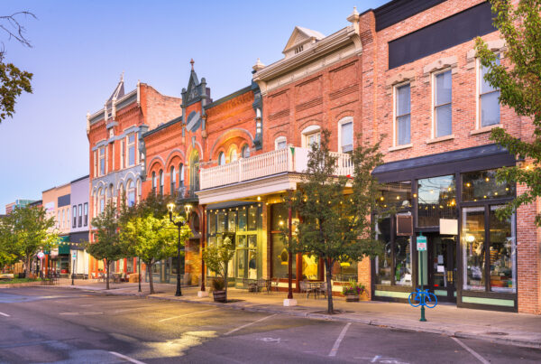 Row of historic brick storefronts with arched windows and outdoor seating along a tree-lined street at dusk.